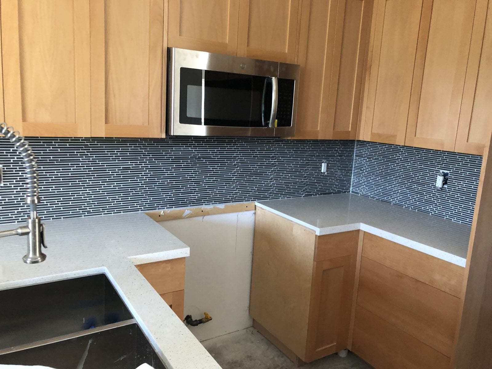 Kitchen with a linear glass mosaic backsplash newly installed above quartz countertops and maple shaker cabinets