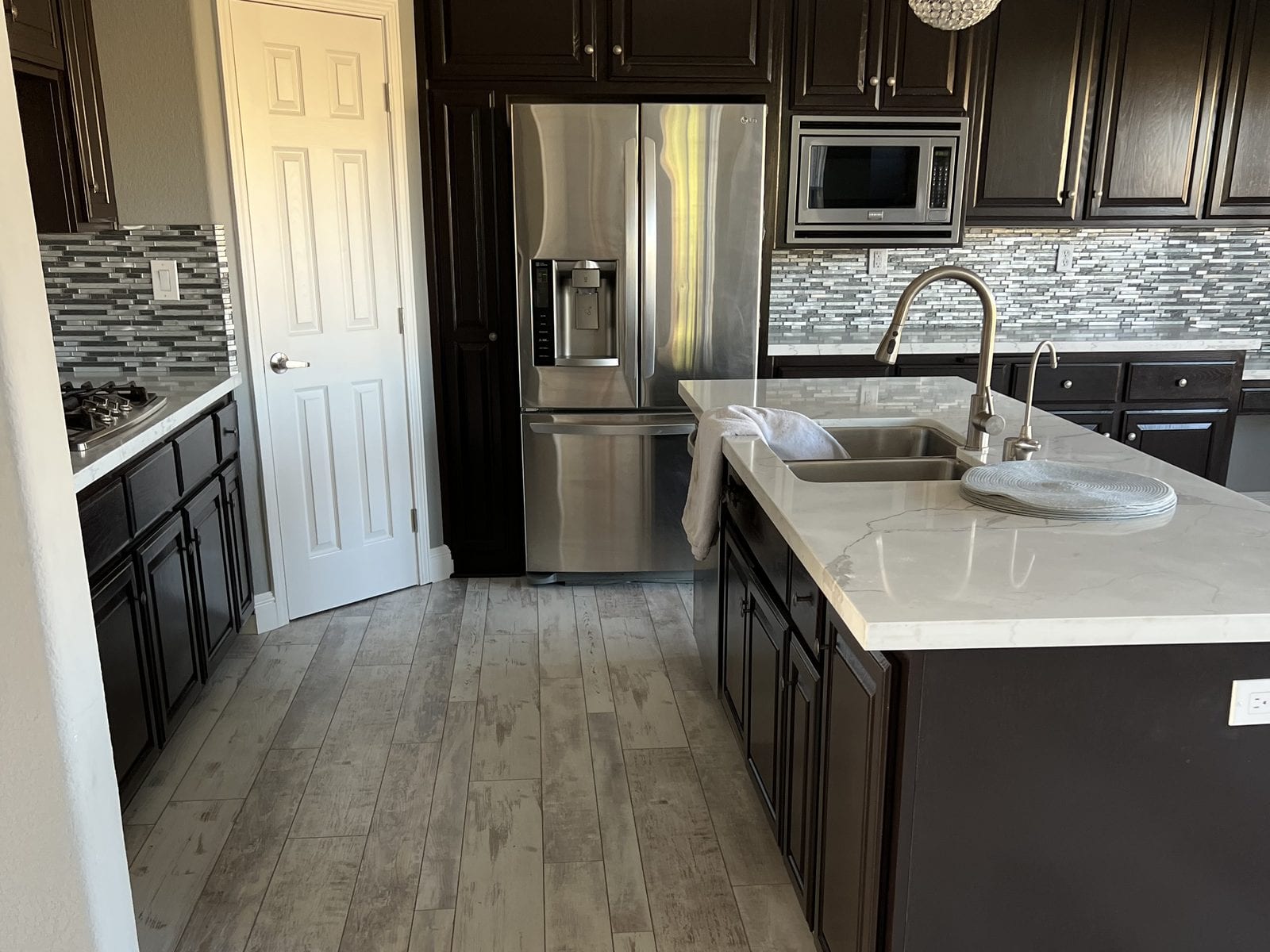 Remodelled kitchen with a quartz-topped island, dark shaker cabinets, stainless appliances and a glass mosaic backsplash over grey wood-look plank flooring