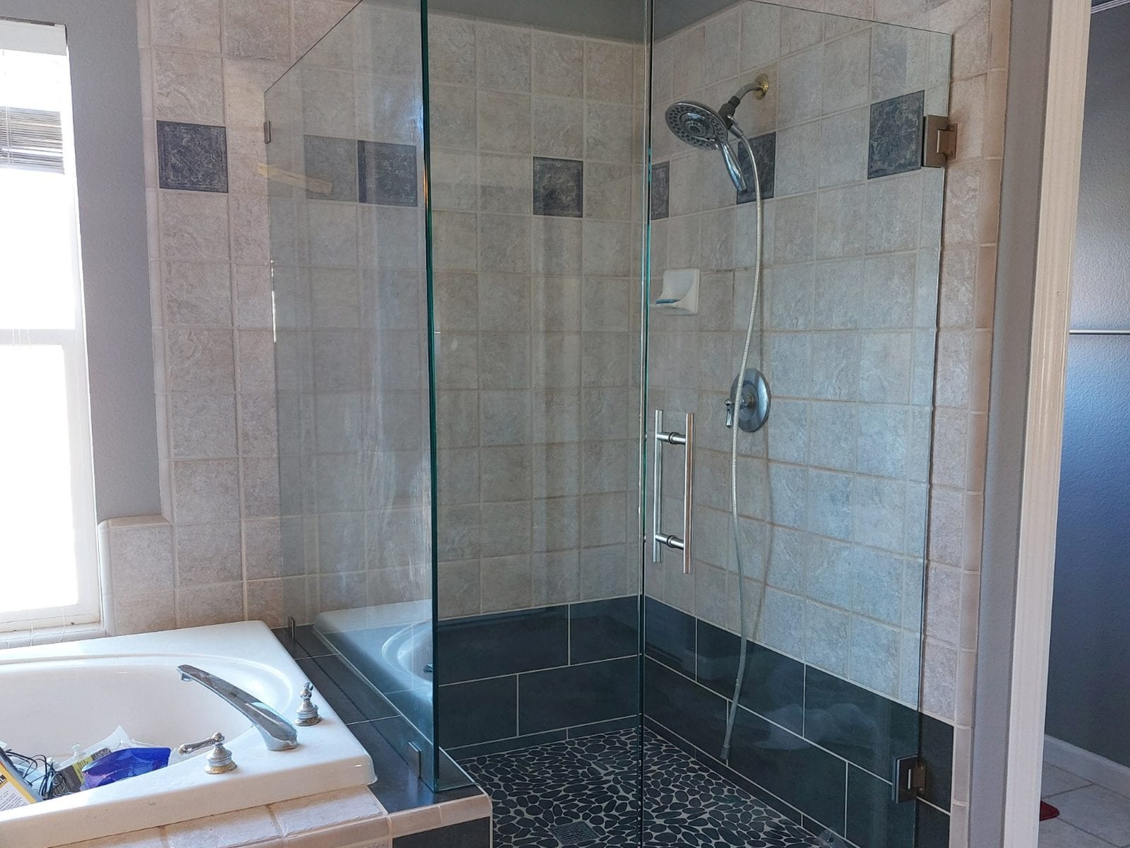 Finished walk-in shower with a frameless glass enclosure, stone-look wall tile, a slate tile band and a black pebble mosaic floor, beside a corner soaking tub
